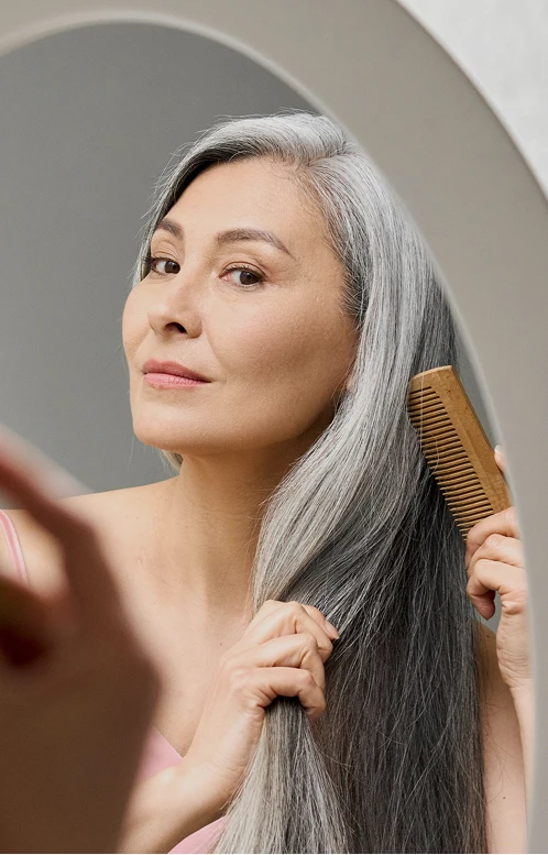A woman with long, elegant grey hair smiles as she uses a wooden comb, showcasing hair health and vitality.