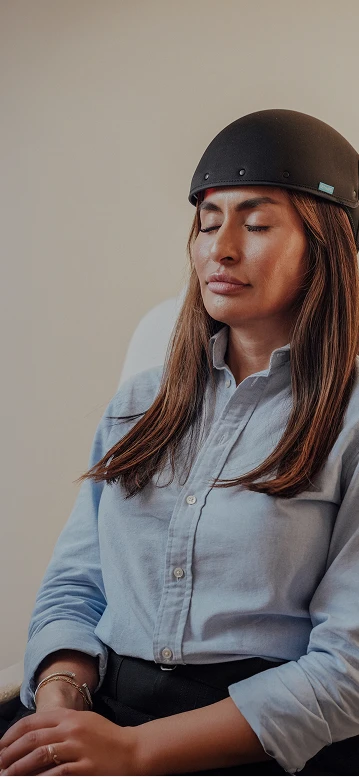 A female patient sits with her eyes closed wearing a black clinical laser cap for a light-based hair regrowth therapy session.