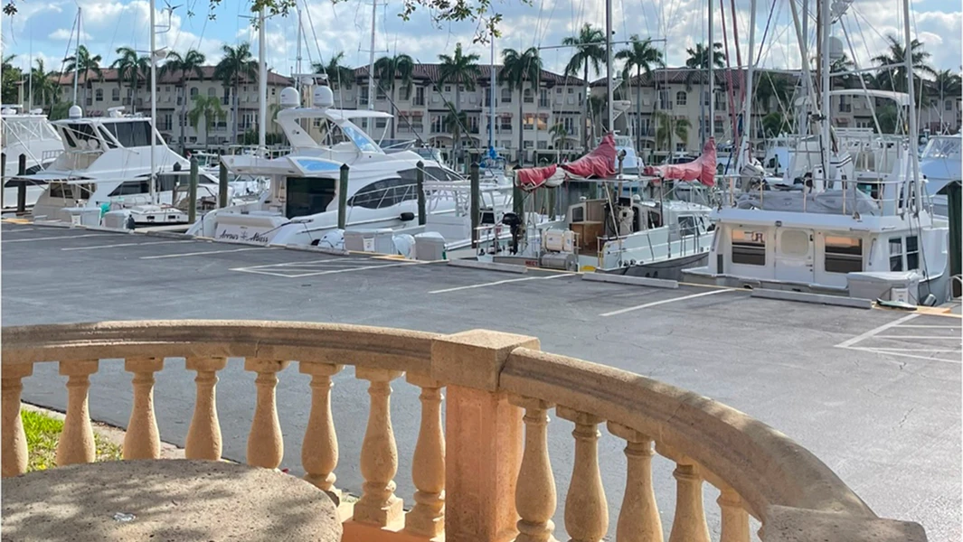 A view from a balcony with stone balusters overlooking a sunny marina filled with yachts and white waterfront buildings.