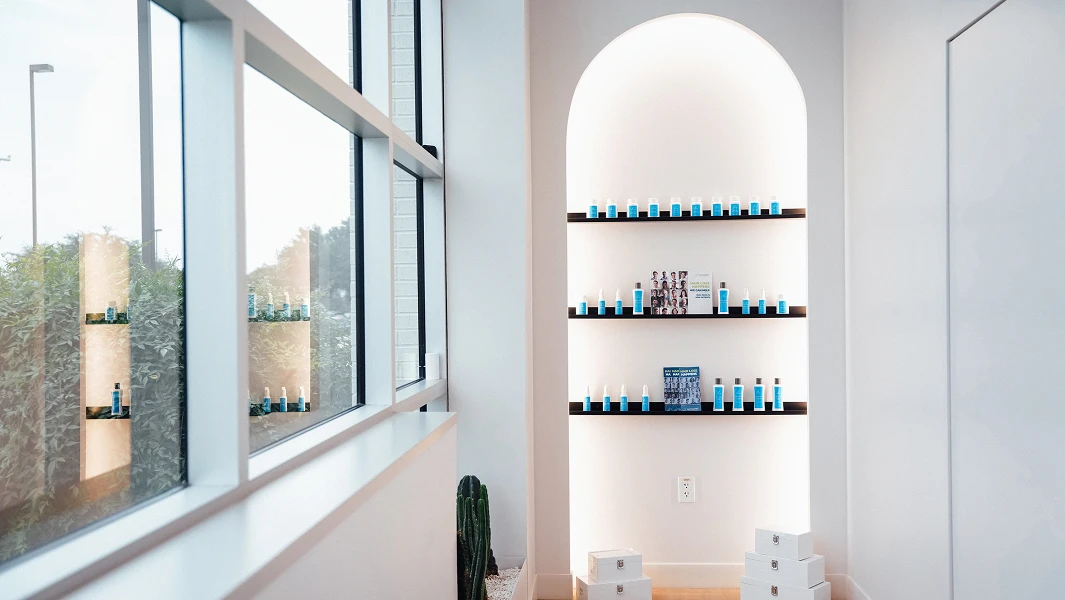 A bright, minimalist clinical interior featuring a large window and a white arched recessed shelf displaying various blue and white professional hair care products.