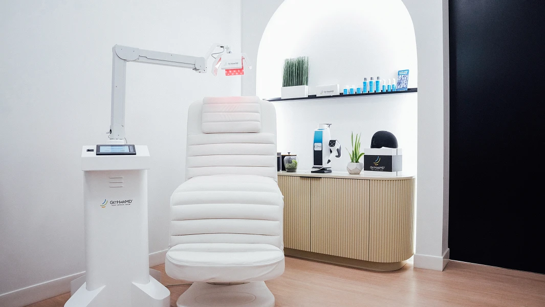 A clinical treatment room featuring a white upholstered medical chair positioned under a specialized red-light therapy arm. Professional hair care products and a laser cap are displayed on a cabinet in the background.