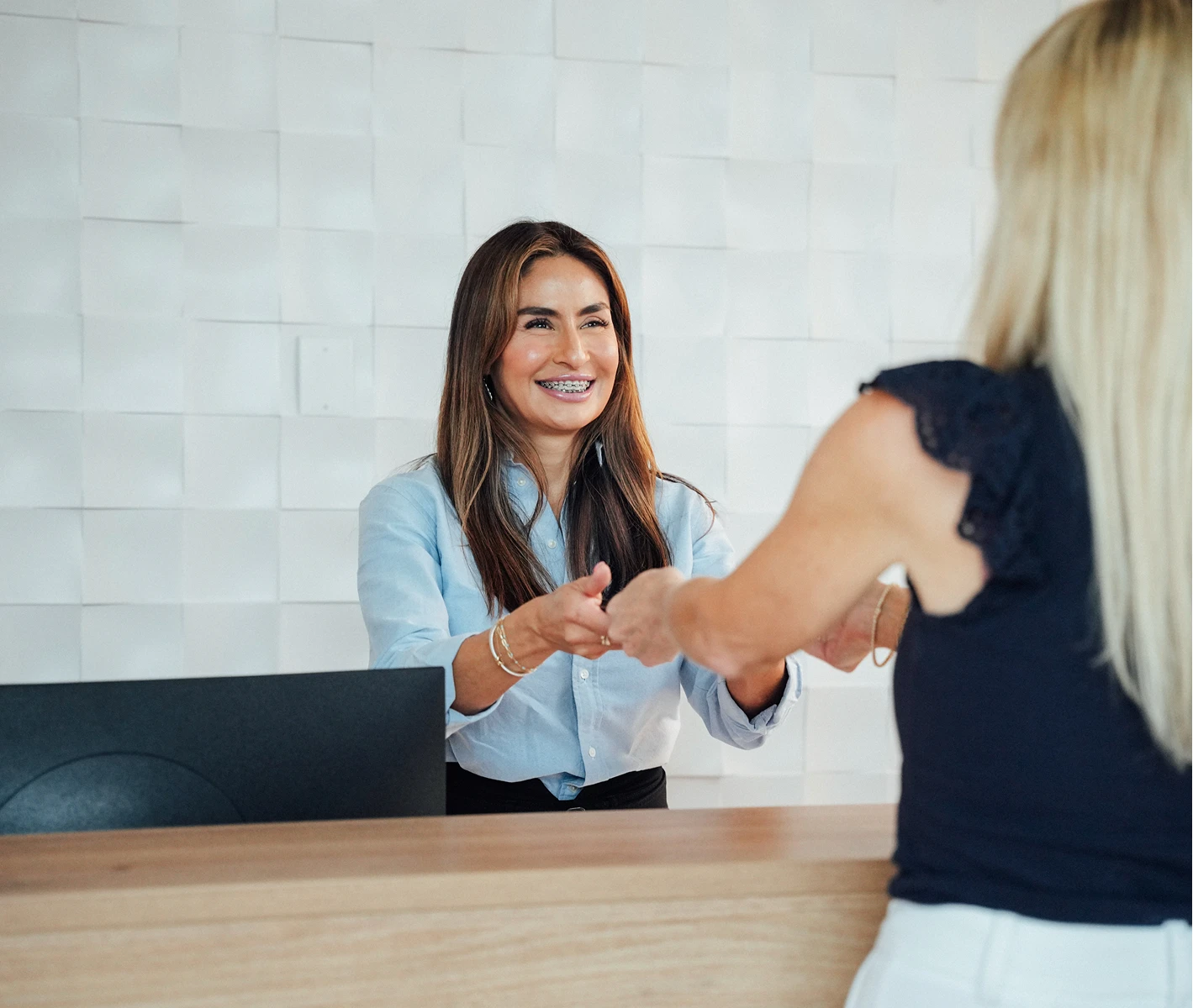 A friendly receptionist with brown hair and braces smiling as she shakes hands with a patient over a wooden reception desk.