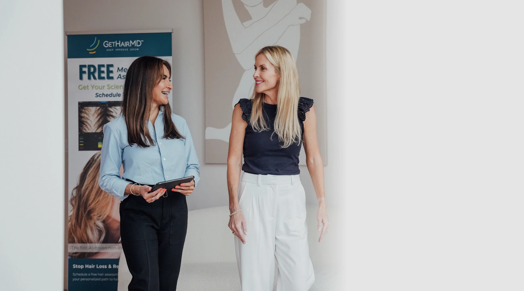 Two women, a clinical staff member holding a tablet and a blonde patient, walking together and chatting in a bright, modern office hallway.