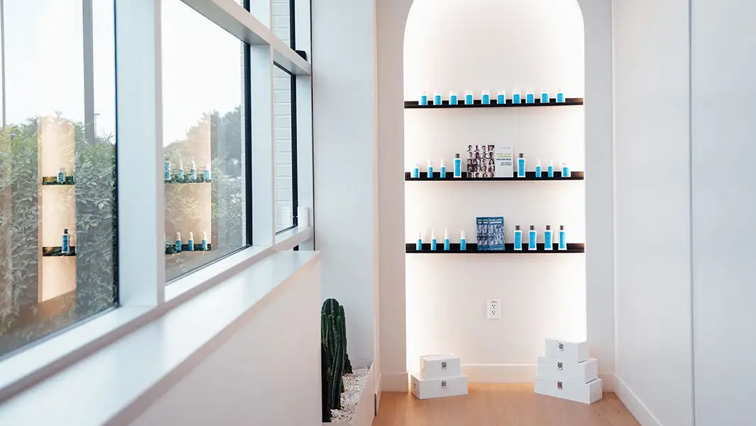 A bright, minimalist clinical interior featuring a large window and a white arched recessed shelf displaying various blue and white professional hair care products.