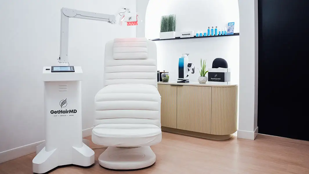 A clinical treatment room featuring a white upholstered medical chair positioned under a specialized red-light therapy arm. Professional hair care products and a laser cap are displayed on a cabinet in the background.