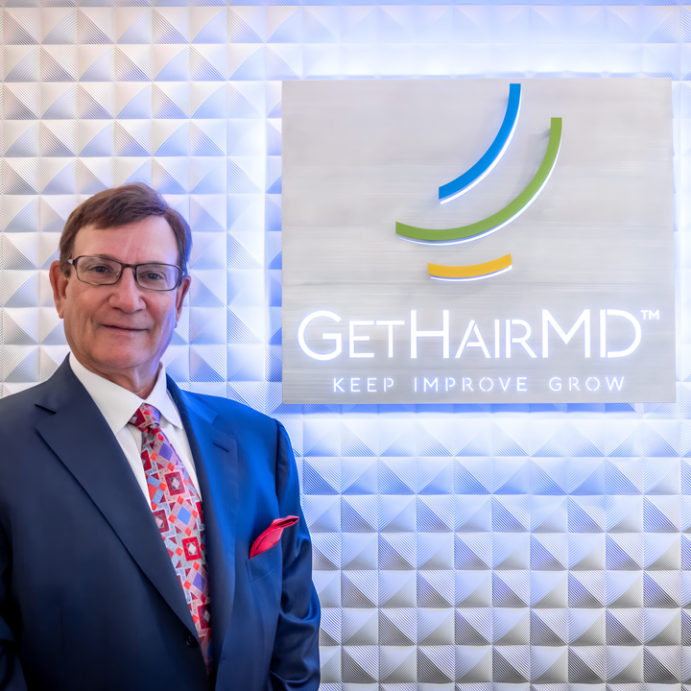 A professional portrait of a male doctor standing proudly in front of a modern, illuminated Get Hair MD "Keep, Improve, Grow" sign.