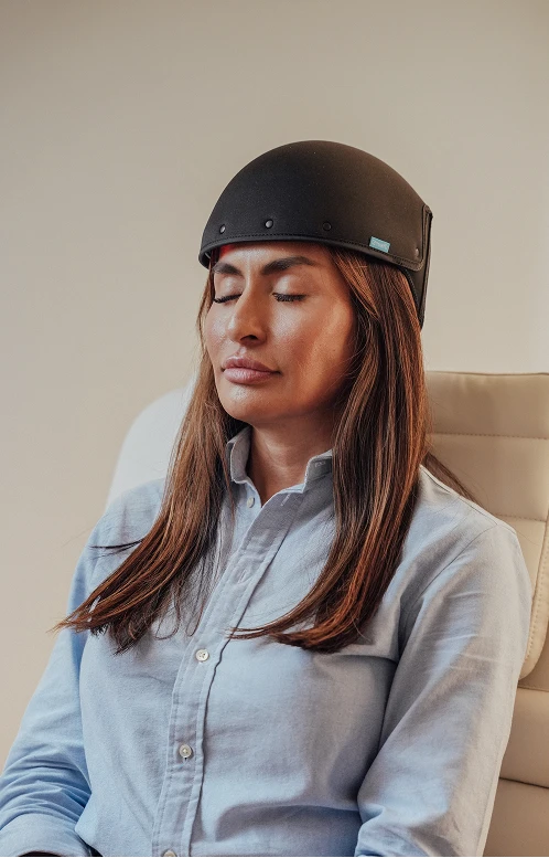 A female patient sits with her eyes closed wearing a black clinical laser cap for a light-based hair regrowth therapy session.