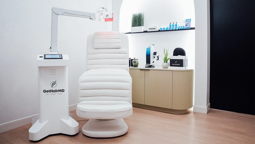 A clinical treatment room featuring a white upholstered medical chair positioned under a specialized red-light therapy arm. Professional hair care products and a laser cap are displayed on a cabinet in the background.