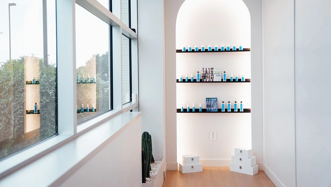 A bright, minimalist clinical interior featuring a large window and a white arched recessed shelf displaying various blue and white professional hair care products.