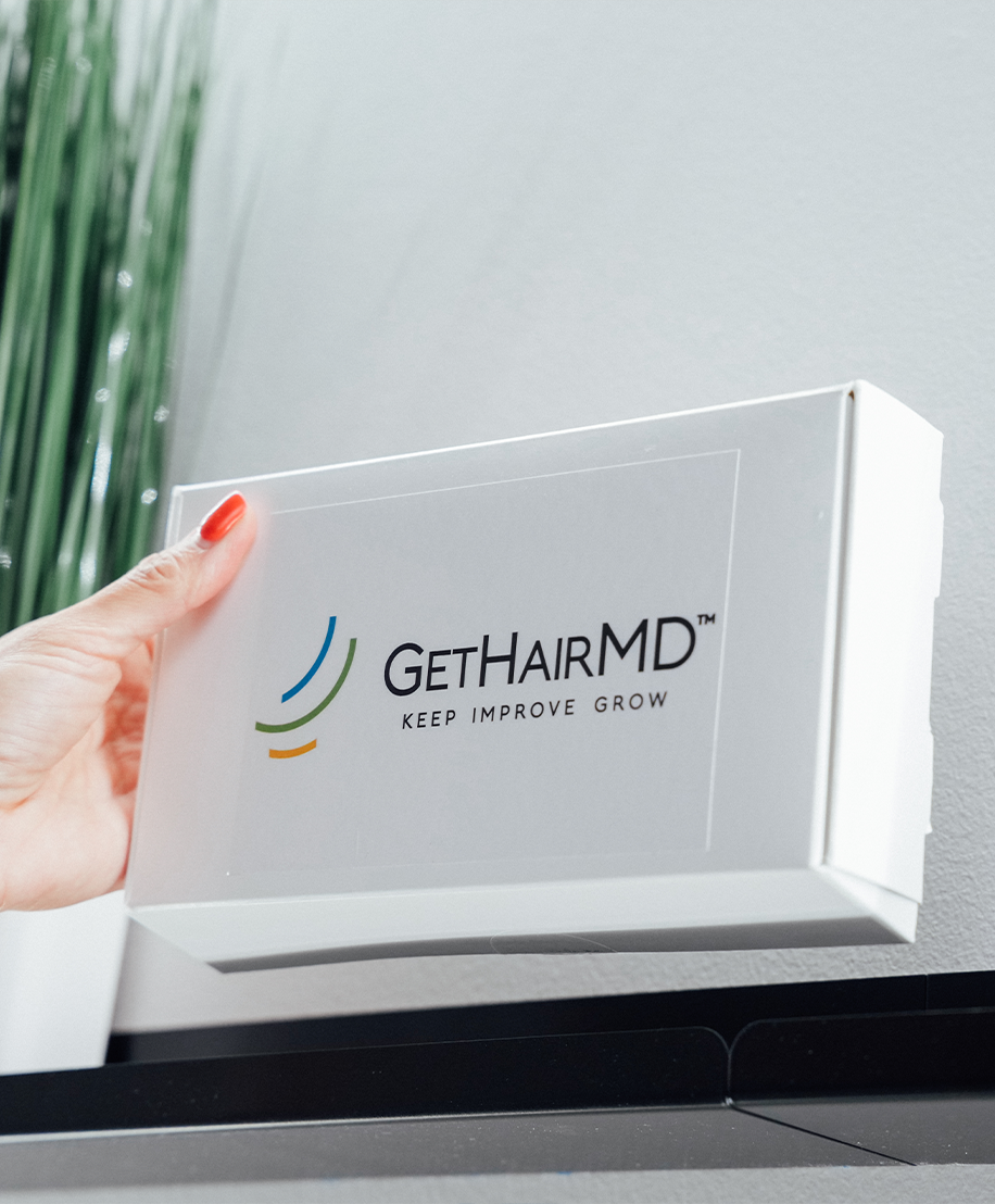 A person's hand holding a white Get Hair MD at-home care kit against a bright wall.
